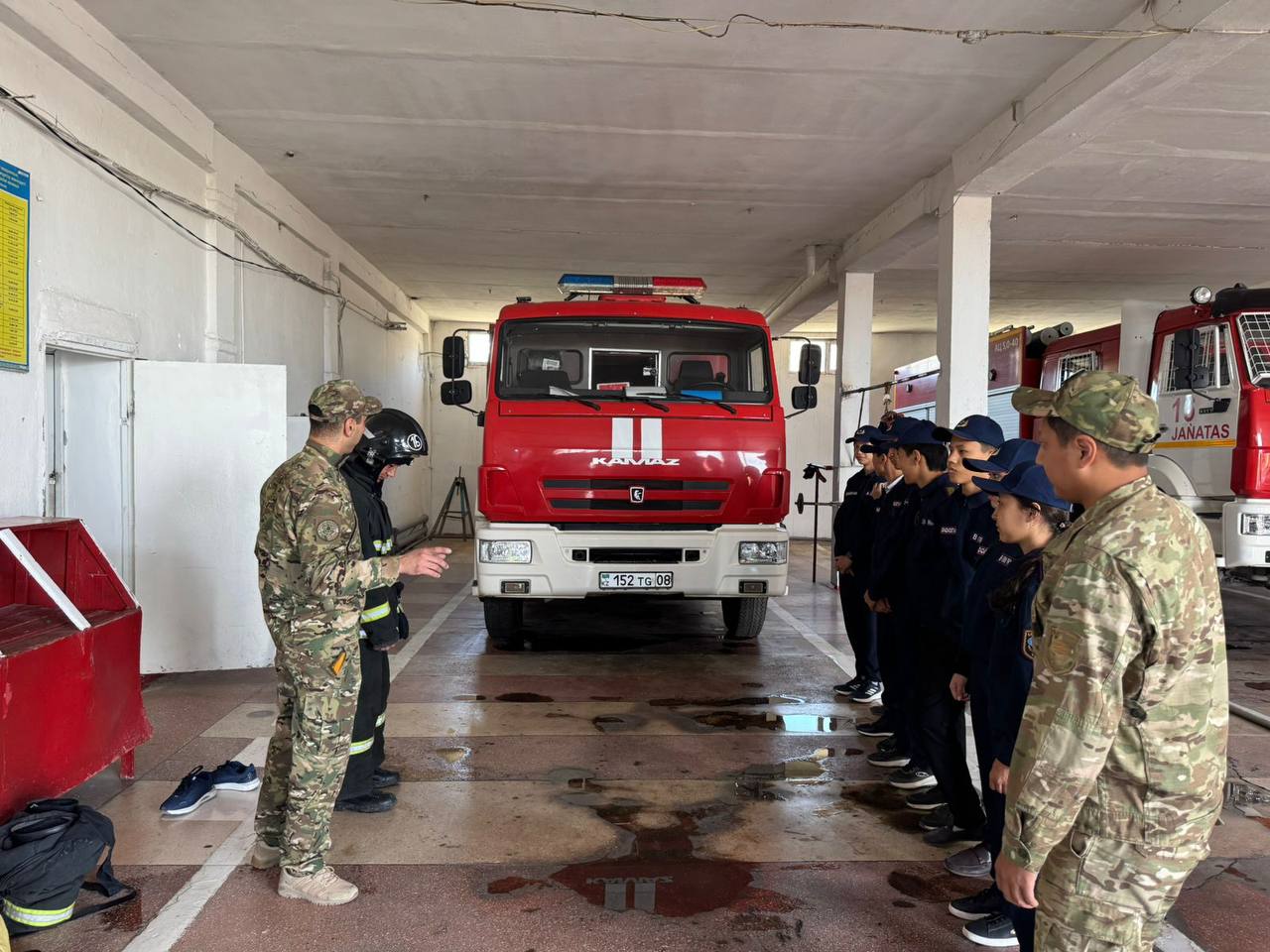 Өрт сөндіру гаражында әскери формадағы адамдар мен мамандардың жиналысы, алда қызыл өрт сөндіру көлігі тұр.