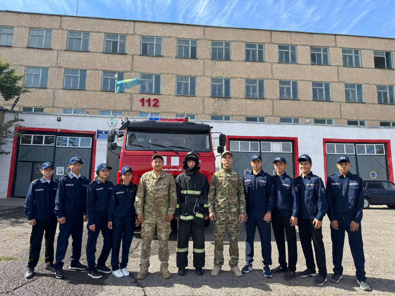 Өрт сөндіру станциясының алдында өрт сөндіру көлігімен бірге тұрған құтқарушылар мен курсанттар тобы.