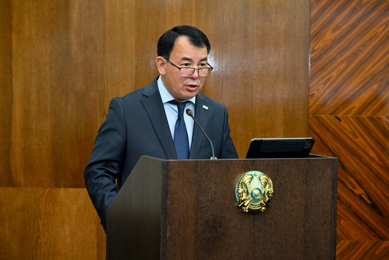 Көзілдірік киген, костюм киген ер адам трибунада сөйлеп тұр.