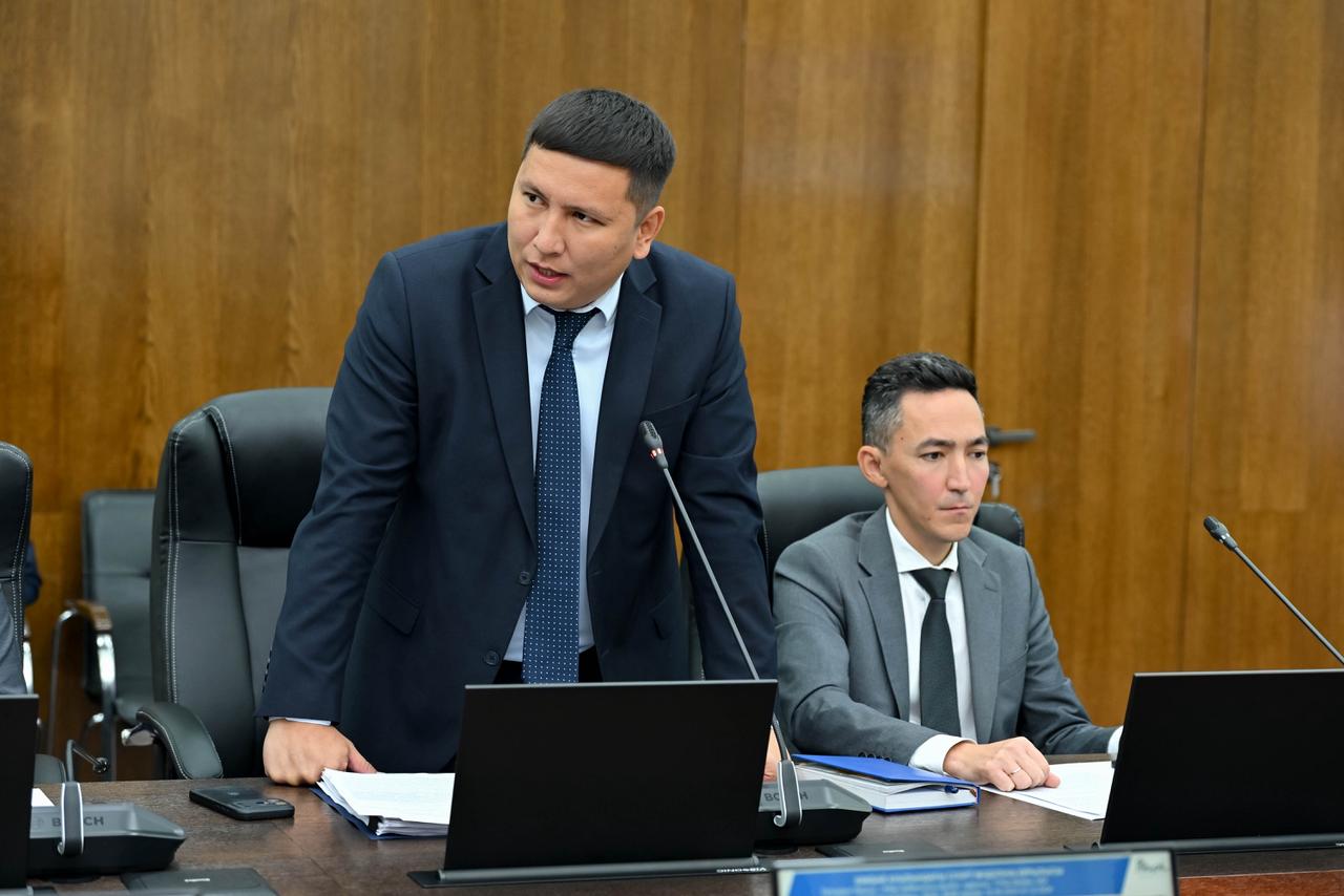 Костюм киген екі ер адам жиналыста отыр, бірі тұрып сөйлеп жатыр.
