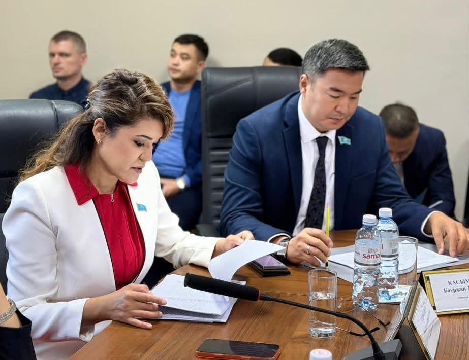 Жыналыста құжаттарды қарап отырған ресми киінген ерлер мен әйел.