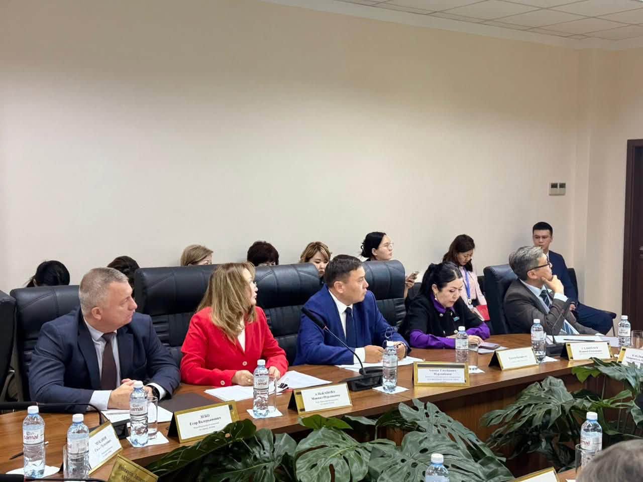 Жиналыс залында үстел басында отырған ресми киінген адамдар тобы.