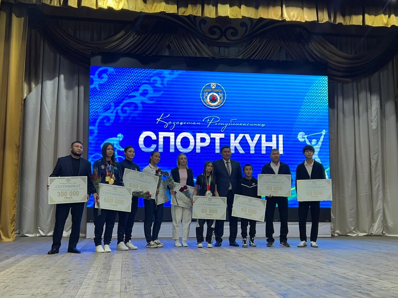 Группа людей с сертификатами на награждении в честь Дня спорта в Казахстане на фоне большого синего экрана.