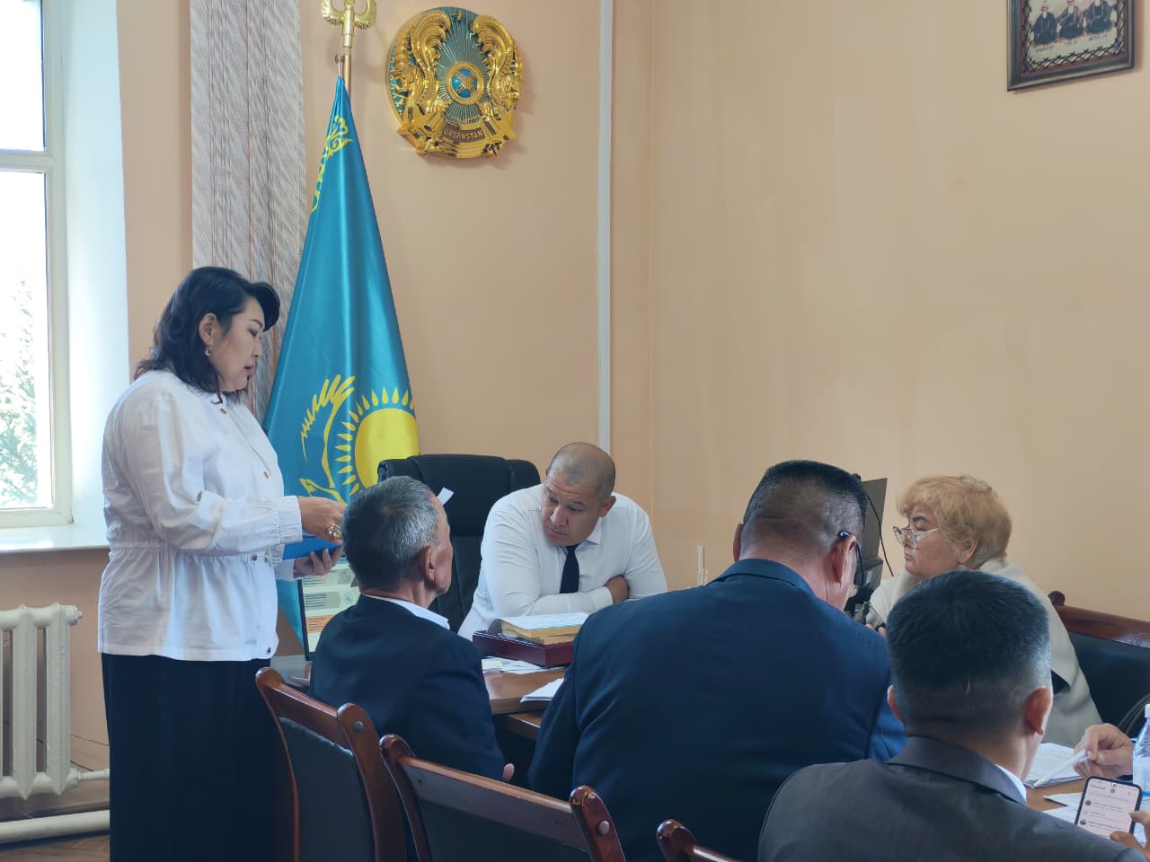Қазақстан Республикасының мемлекеттік рәміздері бар бөлмеде өткізіліп жатқан ресми жиналыс.