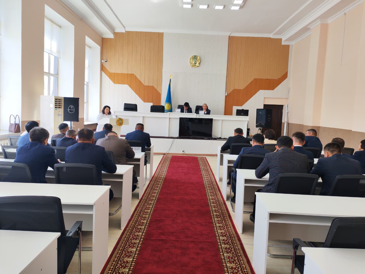 Конференция залындағы мәслихат сессиясының отырысында : спикер сөйлеп тұр, ал қатысушылар үстелдерде отыр.