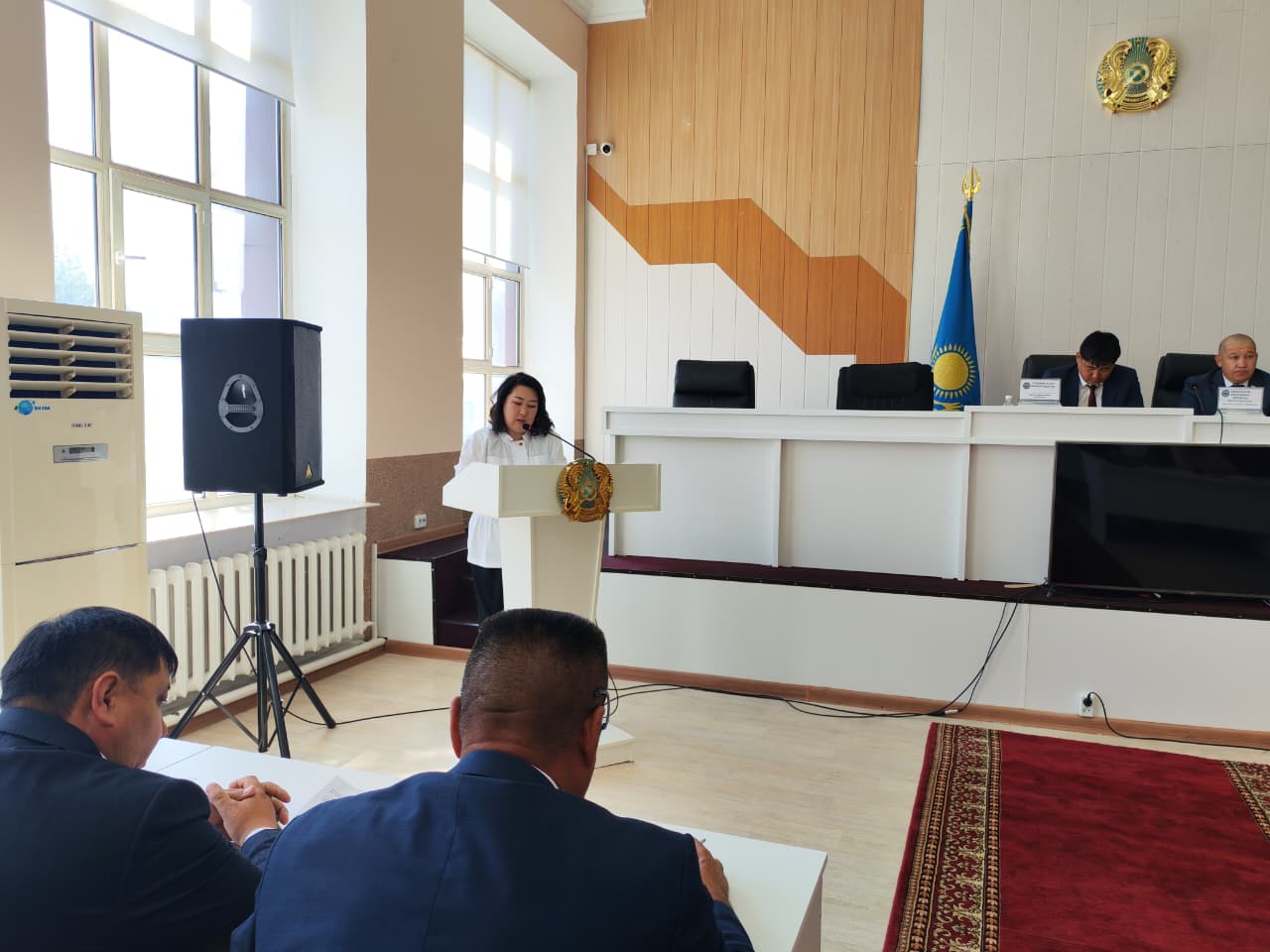 Трибунада сөйлеп тұрған әйел және алдында отырған депутаттар