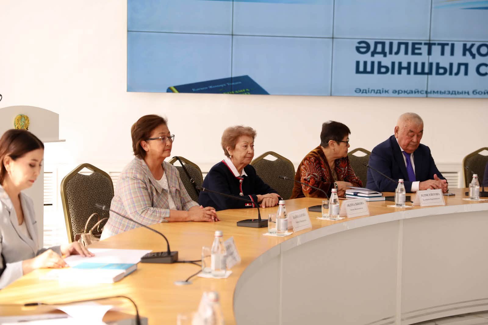 Жиналыс үстелінде отырған төрт адам. Фонда қазақ тілінде мәтіні бар экран көрінеді.