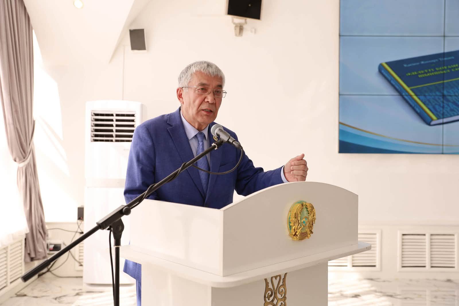Көк костюм киген ер адам трибунада микрофонмен сөйлеп тұр.