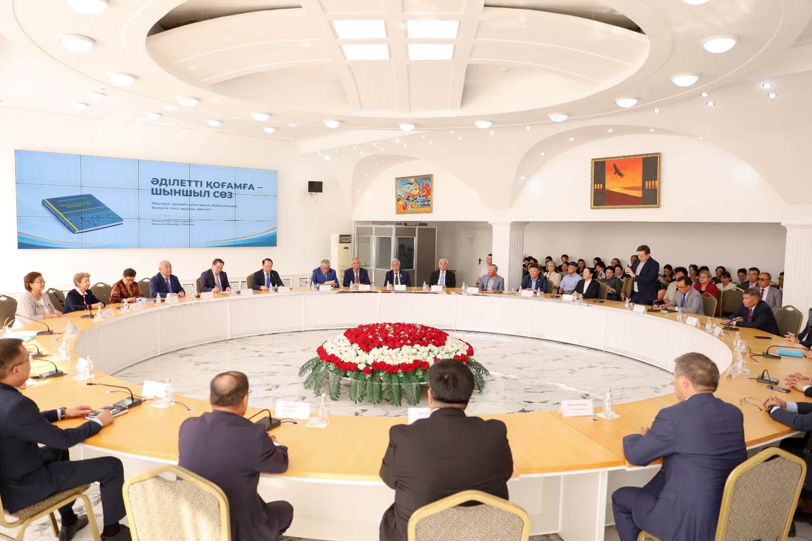 Дөңгелек үстелде жиналған ресми адамдар және алдындағы презентация экраны.