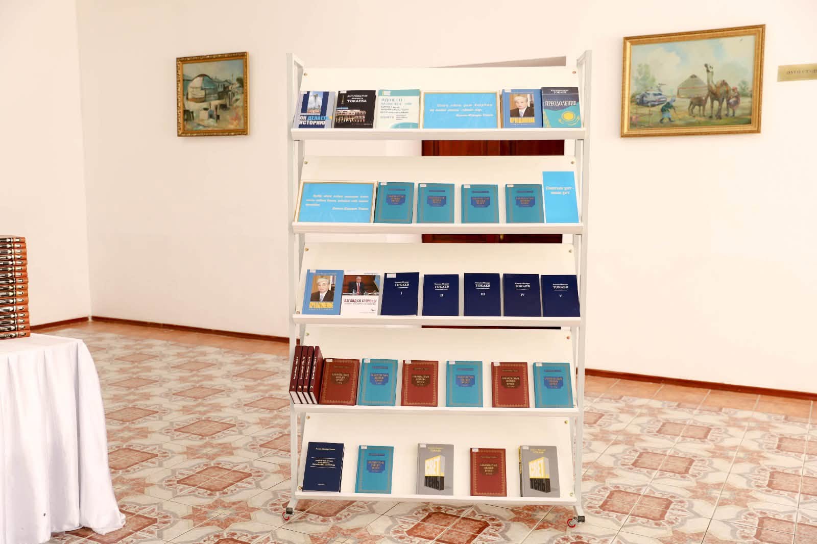 Кітаптар қойылған ақ түсті ашық сөре. Сөренің артында қабырғаға ілінген екі картина бар.