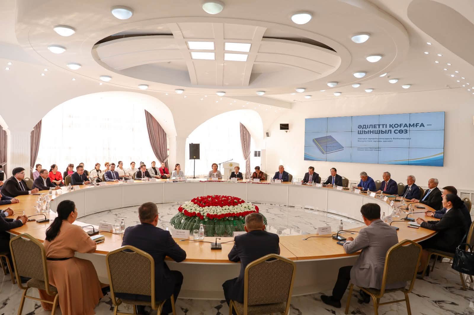 Дөңгелек үстелде жиналған ресми адамдар тобы және презентация экраны.