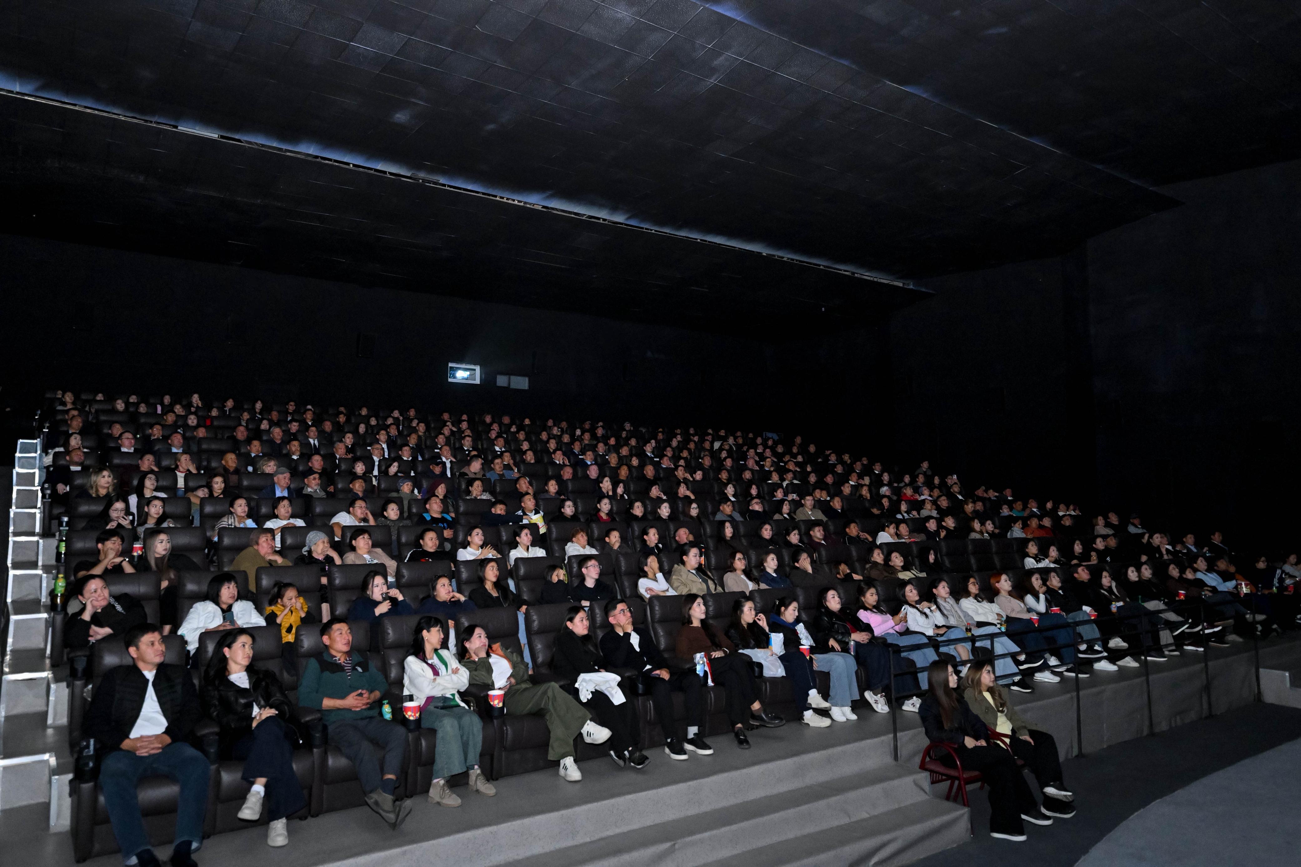 Кинотеатр залында экранға қарап отырған көрермендер тобы.