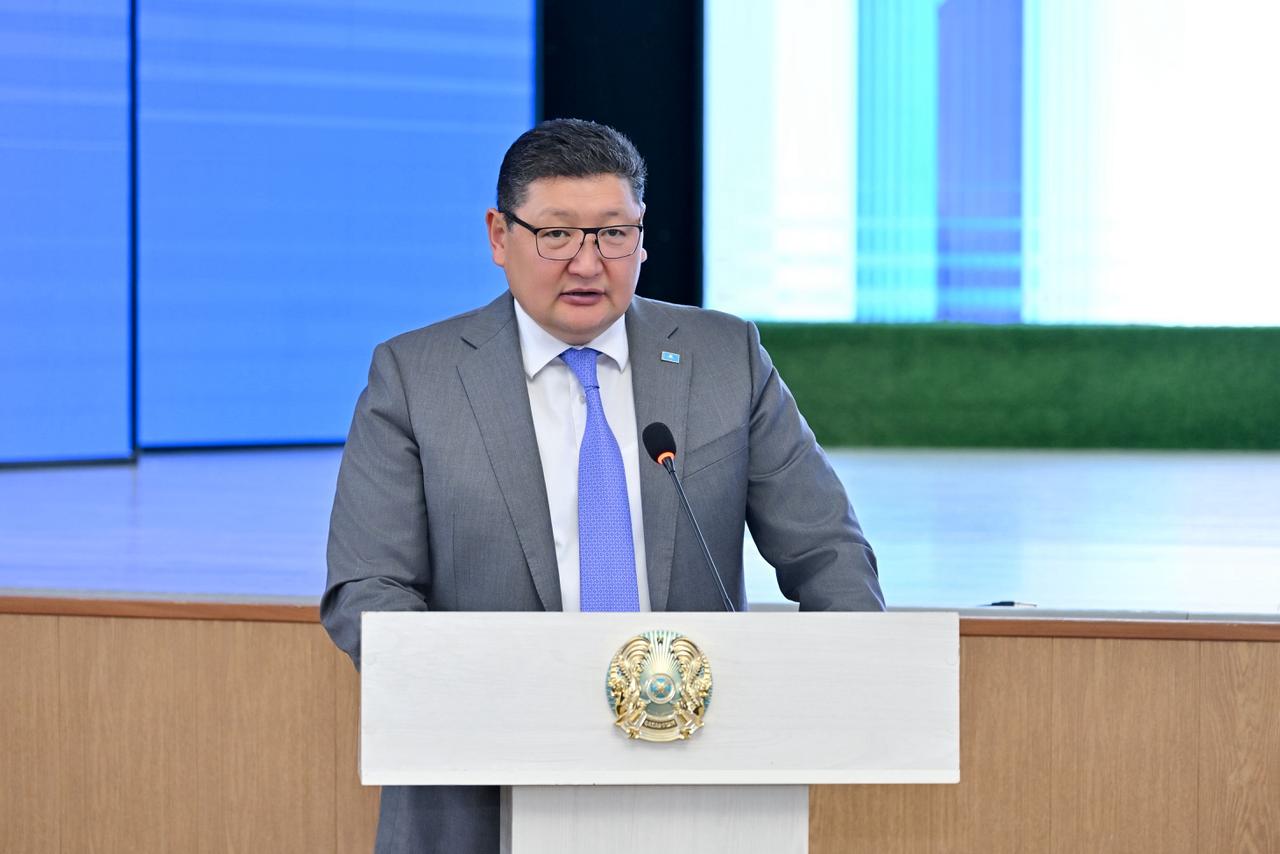 Сұрақ қою трибунасында тұрған сұр костюмді, көзілдірікті ер адам.