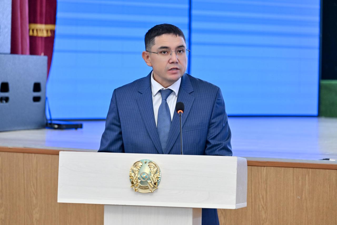 Көк костюм киген ер адам трибунада сөйлеп тұр.