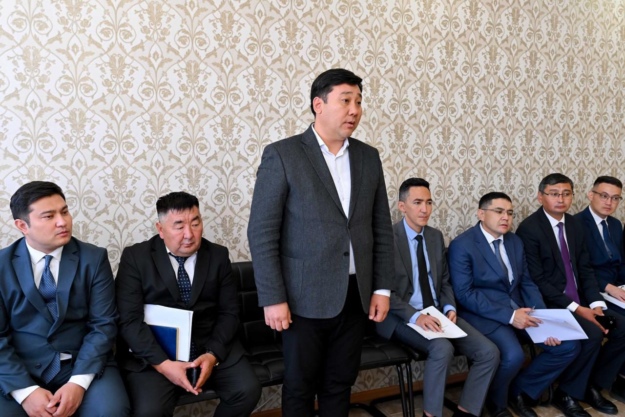 Костюм киген ер адамдар тобының жиналысы. Ортада тұрған ер адам сөйлеп жатыр.