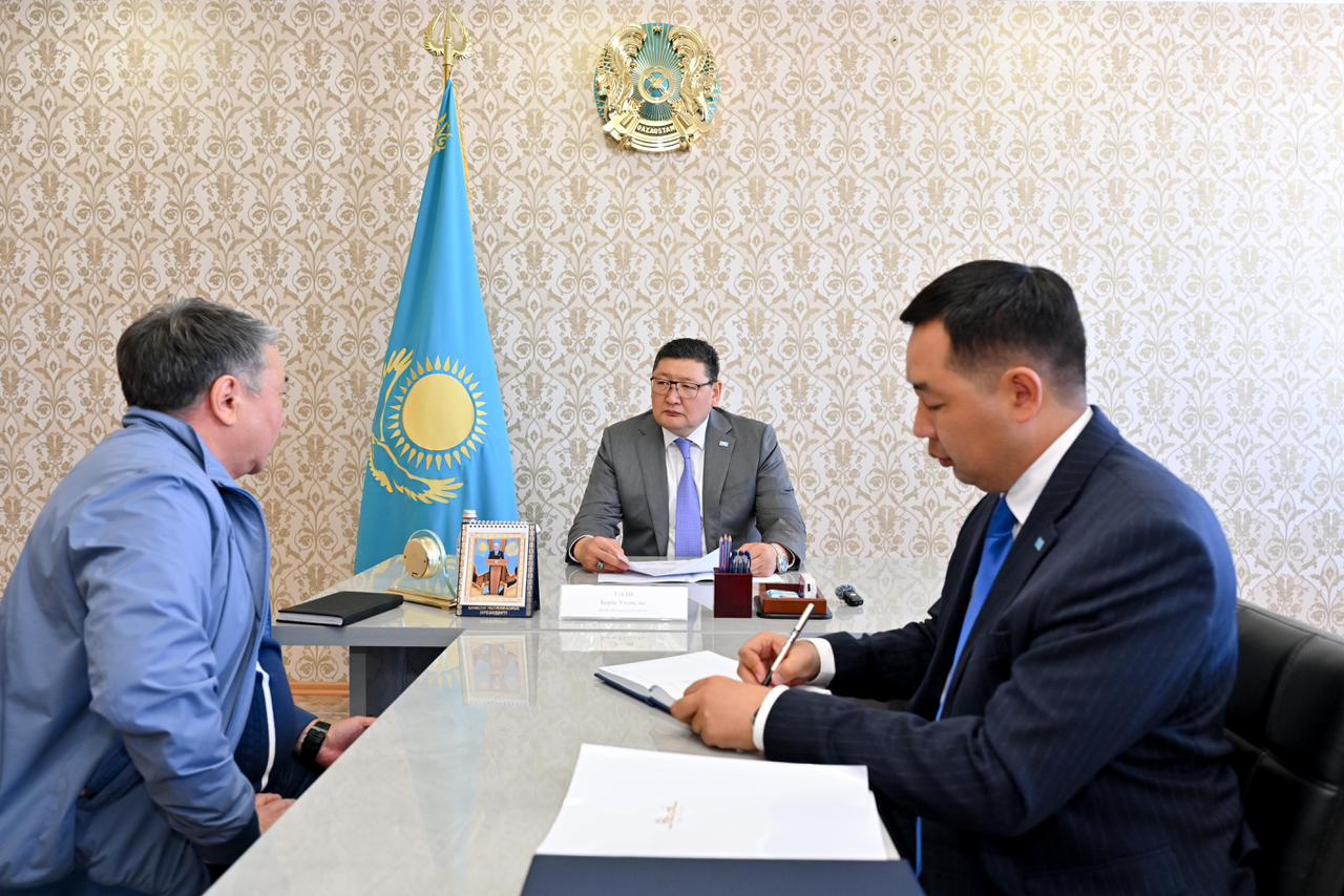 Қазақстан Республикасының мемлекеттік рәміздері бар кеңседе үш ер адамның жұмыс кездесуі.