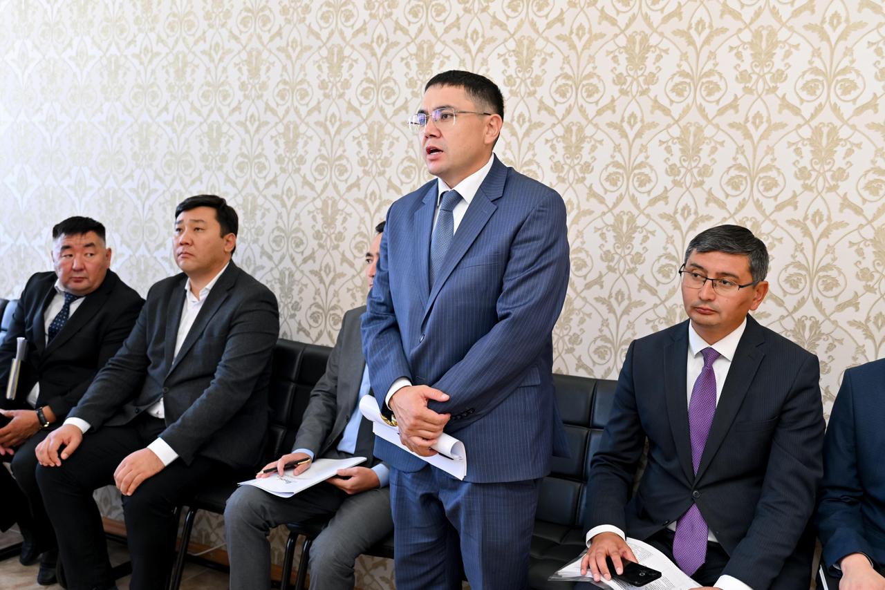 Көк костюм киген ер адам жиналыста сөйлеп тұр, ал жанында басқа ерлер отыр.