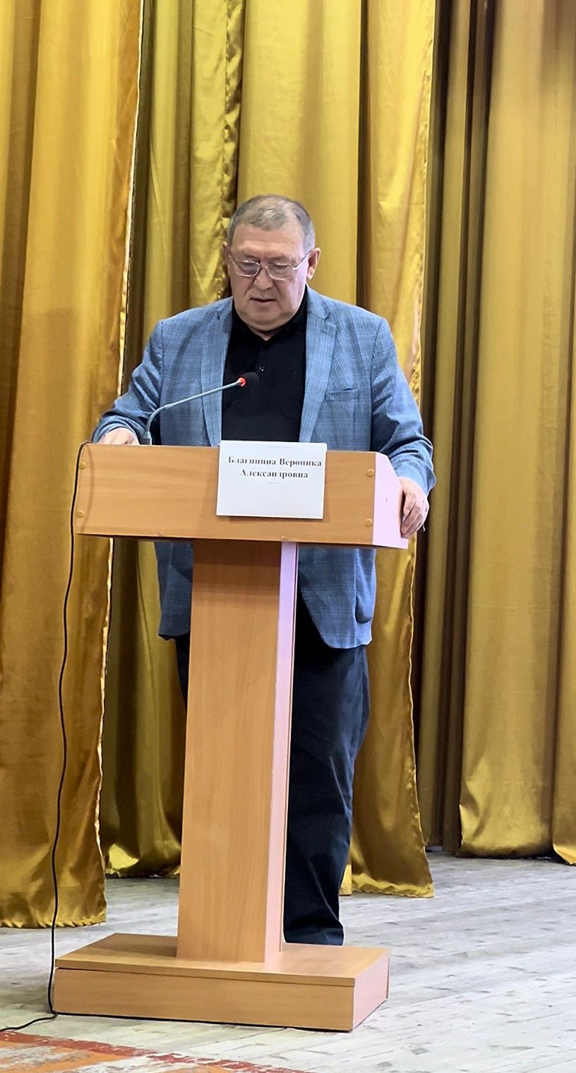 Алтын түсті перде алдындағы трибунада сөйлеп тұрған көзілдірікті ер адам.