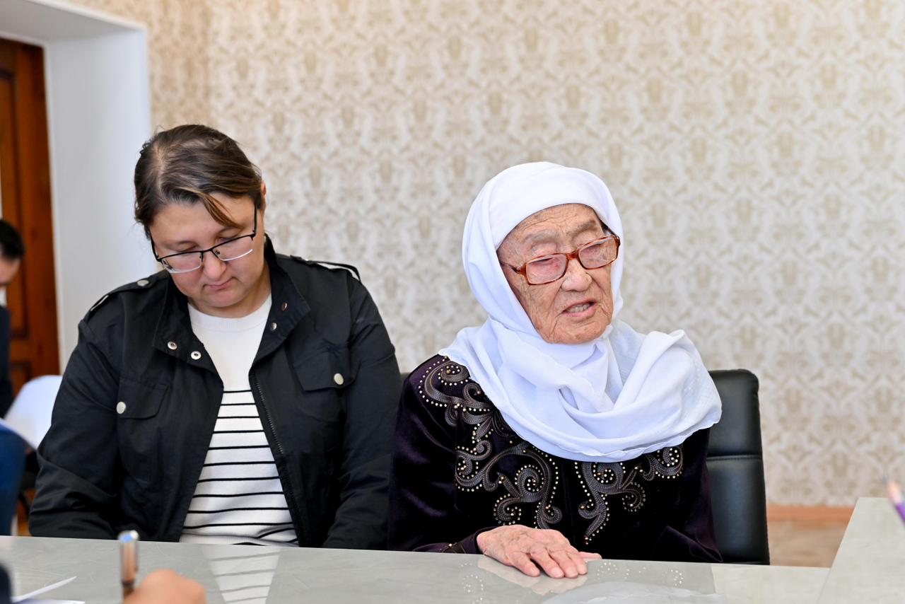 Көзілдірік таққан екі әйел: жас әйел және ақ жаулықты қарт әйел үстел басында отыр.