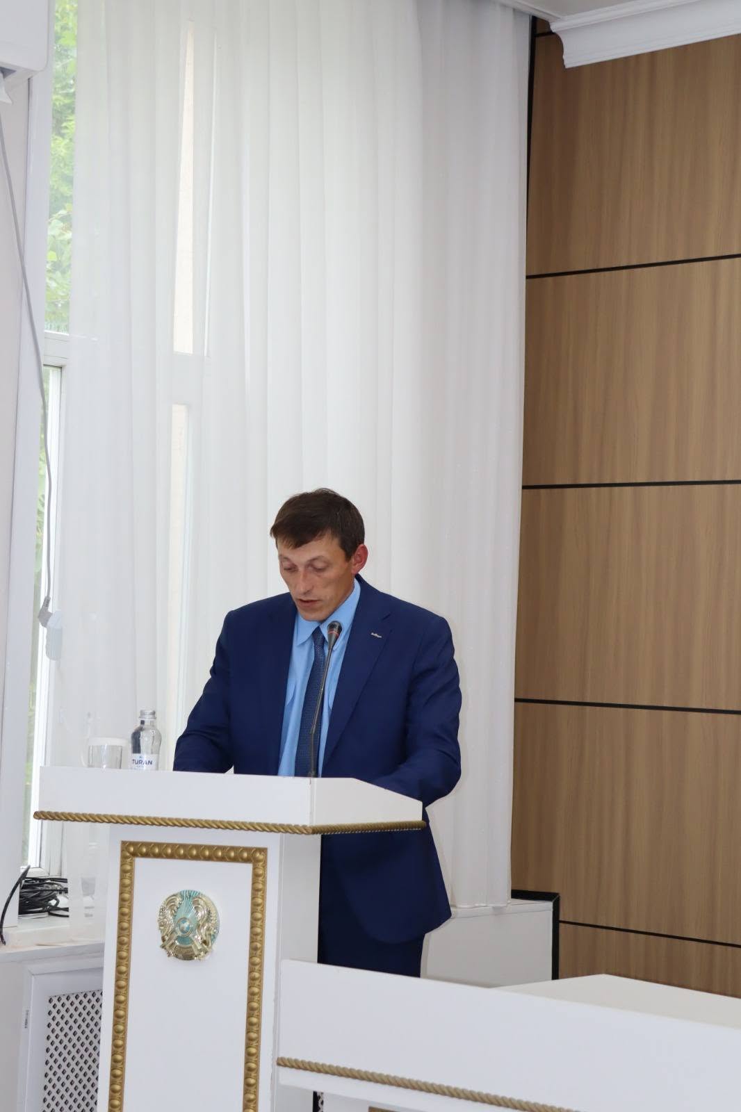 Көк костюм киген ер адам трибунада сөйлеп тұр.