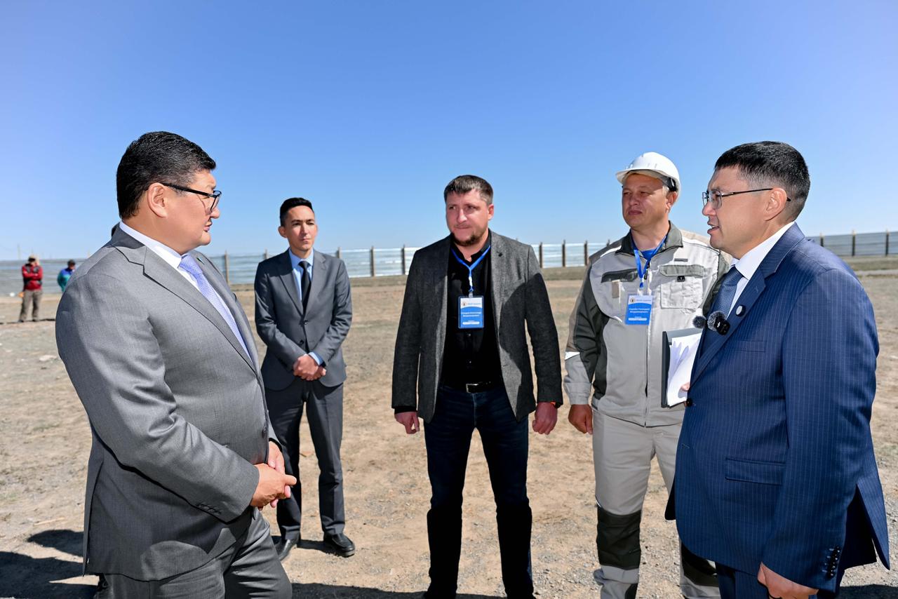 Ашық аспан астында бизнес-костюмдегі және жұмыс киіміндегі ер адамдардың тобы сөйлесіп тұр.