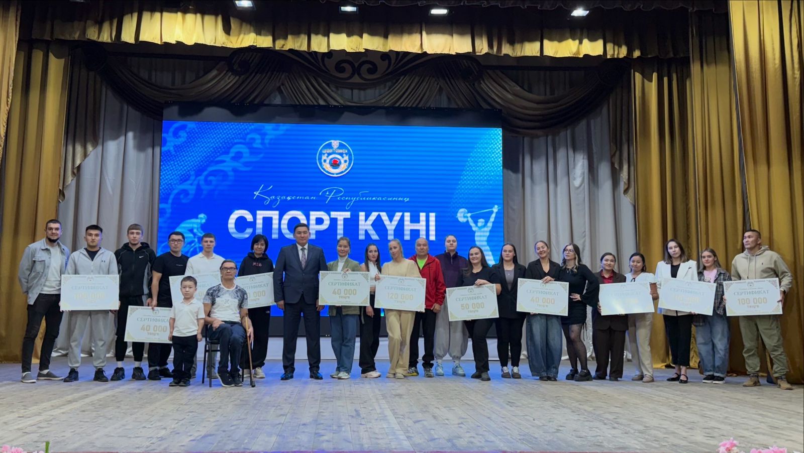 Сахнада «СПОРТ КҮНІ» деп жазылған көк экранның алдында сертификаттары бар адамдар тобы.