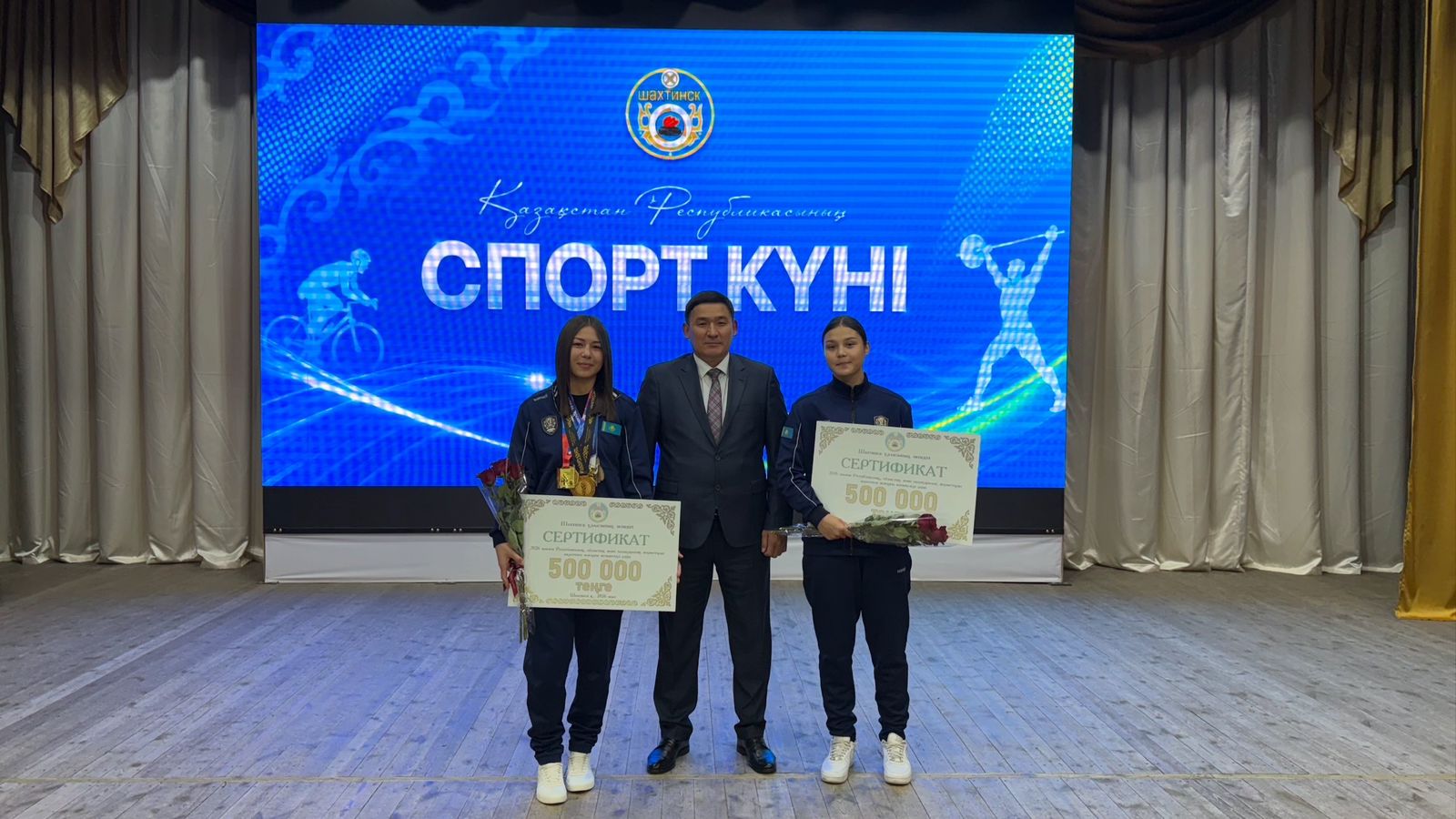 Сахнада «СПОРТ КҮНІ» деп жазылған көк экранның алдында сертификаттары бар адамдар тобы.