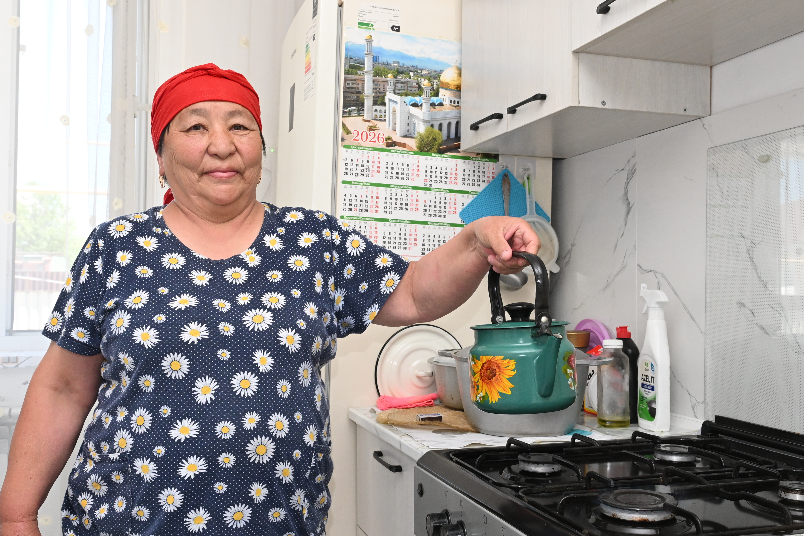 Женщина в красном платке и синем платье в ромашках стоит на кухне и держит за ручку зеленый чайник.