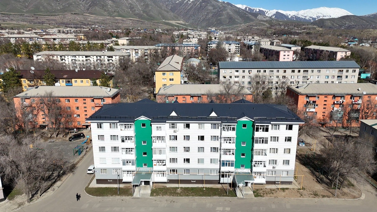 Современный многоквартирный дом с зелеными акцентами на фоне городского массива и заснеженных гор.